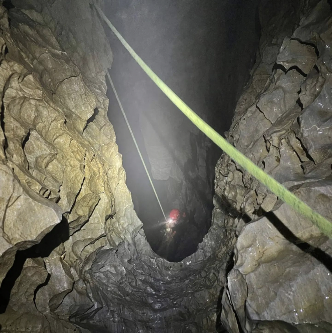 This image shows a person with a red helmet and a head lamp lowering themselves into a big cavern on a green rope. This person is caving in a cave system near Port Alberni on Vancouver Island.