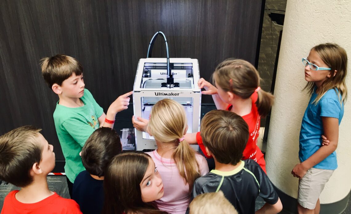 6 kids surrounding a white, square machine. some are pointing at it, some are just looking at it. theres a tube coming out from the top of the box, and the box says "ultimaker"