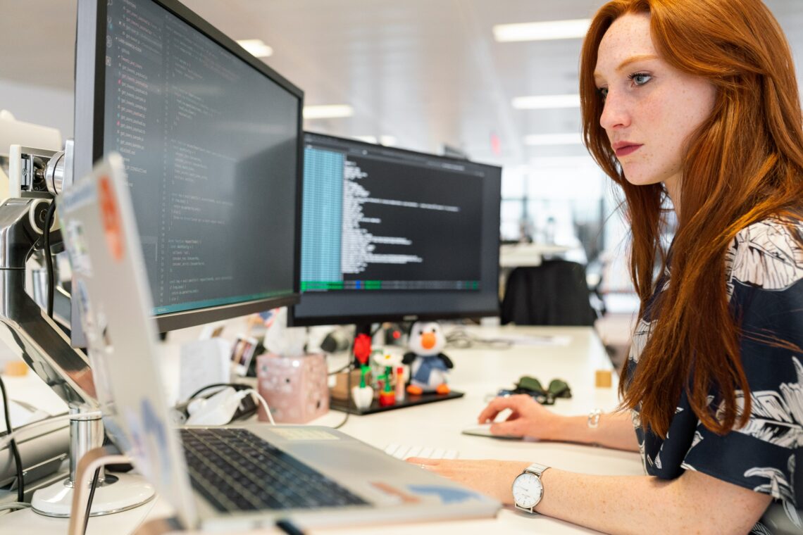 A girl with red hair is looking at two computer moniters and a laptop. She has a focused face and looks like she is doing some coding on the computer monitor on her right.
